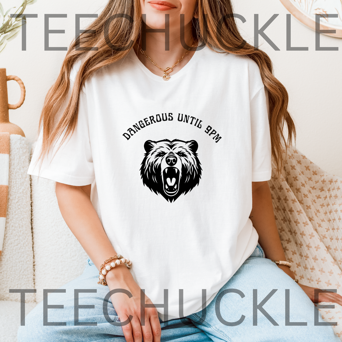 A woman wearing a white short-sleeve graphic T-shirt with a black bear head illustration and the text 'DANGEROUS UNTIL 9PM' across the chest, paired with light blue jeans in a cozy indoor setting.