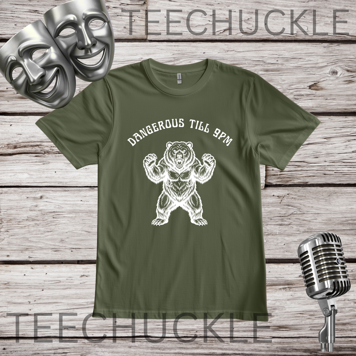 An green crewneck t-shirt with a white graphic of a bear flexing and the text 'DANGEROUS TILL 9PM' printed on the front. The shirt is displayed flat on a rustic wood background with decorative props including two silver comedy masks, a vintage microphone.