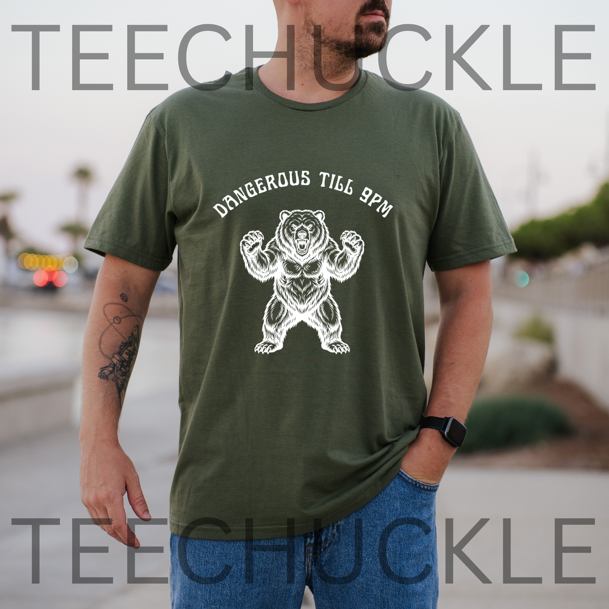 A man wearing an green crewneck t-shirt with a white graphic of a flexing bear and the text 'DANGEROUS TILL 9PM'. and the background is a blurred outdoor street scene.