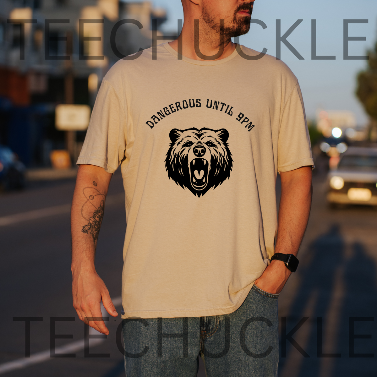 A man wearing a sand graphic t-shirt with a black bear design and the text 'DANGEROUS UNTIL 8PM' on the front, standing outdoors on a street at sunset. 