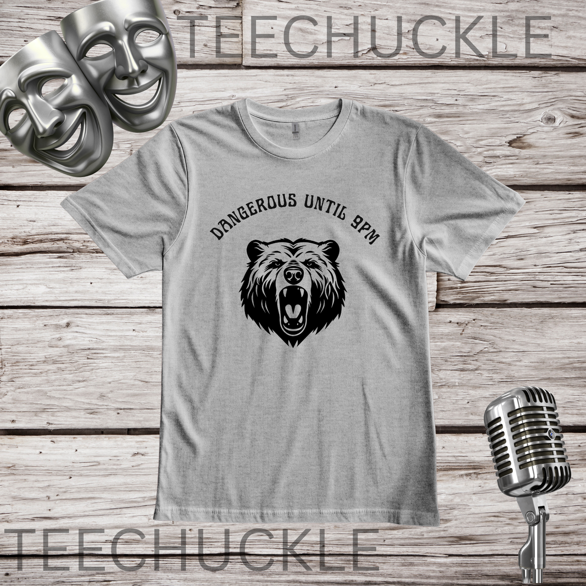 A light gray graphic t-shirt featuring a black bear head illustration and the text 'DANGEROUS UNTIL 9PM' on the front, displayed on a rustic wood plank background with decorative theater masks and a vintage microphone.