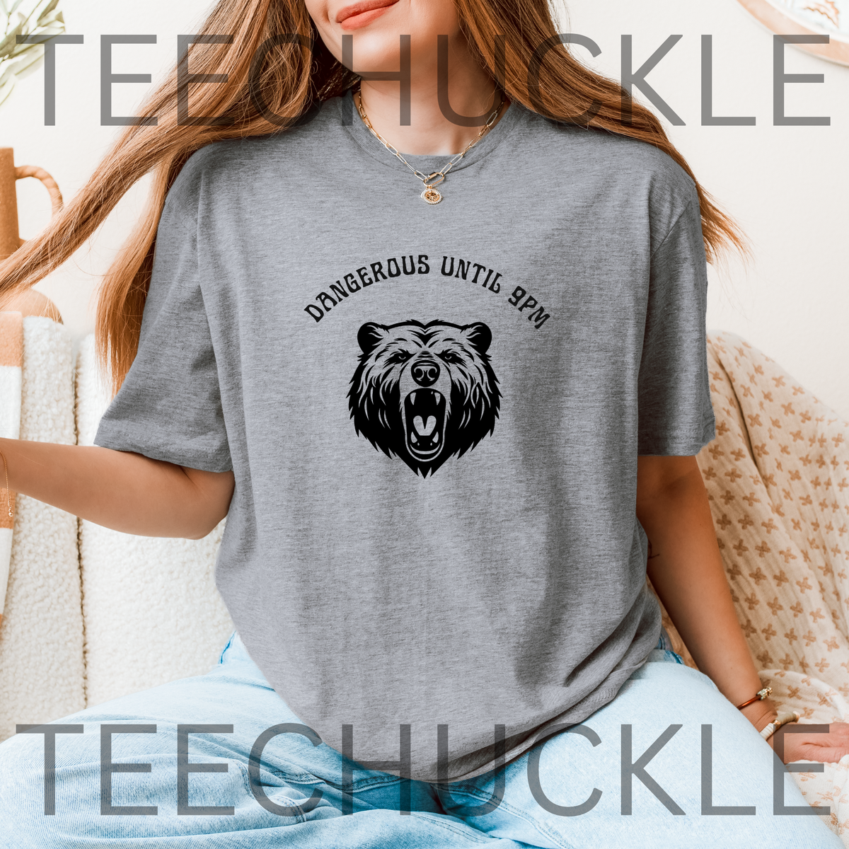 A woman wearing a heather gray short-sleeve graphic T-shirt with a black bear head illustration and the text 'DANGEROUS UNTIL 9PM' across the chest, seated indoors on a textured chair. 