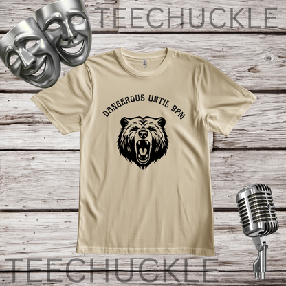 A sand graphic t-shirt featuring a black bear head illustration and the text 'DANGEROUS UNTIL 9PM' on the front, displayed on a rustic wood background with silver comedy masks, a vintage microphone