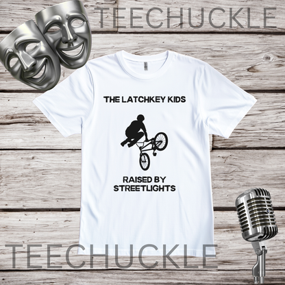 A white graphic t-shirt displayed on a rustic wooden background, featuring a black print of a BMX rider and the text 'THE LATCHKEY KIDS RAISED BY STREETLIGHTS'. The shirt is styled with silver comedy masks and a vintage microphone.