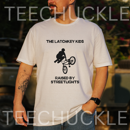 A man wearing a white graphic t-shirt with black text reading 'THE LATCHKEY KIDS RAISED BY STREETLIGHTS' and a bicycle silhouette, photographed outdoors with a blurred background. 