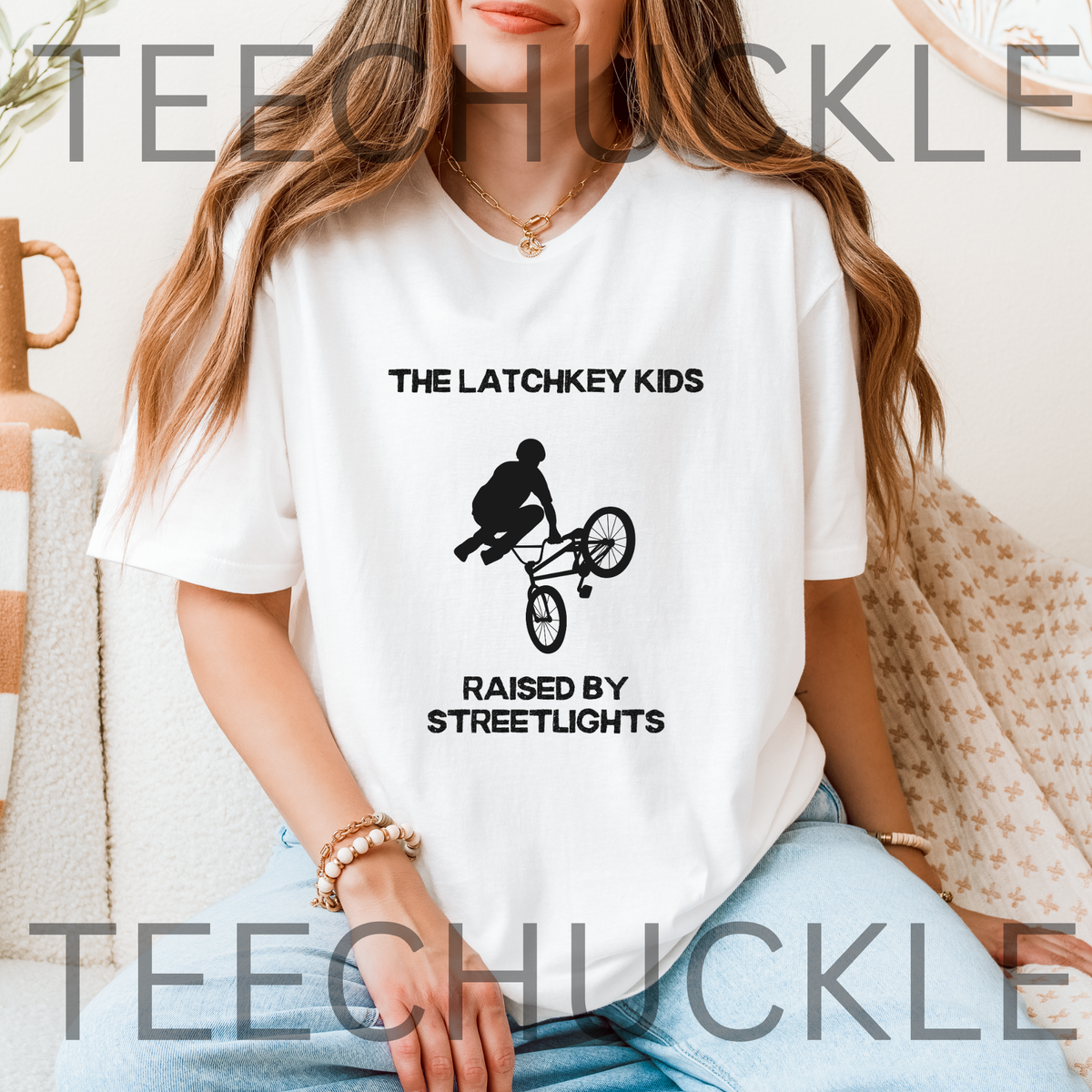 A woman wearing a white graphic t-shirt with a black print that reads 'THE LATCHKEY KIDS RAISED BY STREETLIGHTS' with a BMX rider silhouette, sitting on a couch.