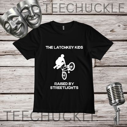 A black short-sleeve graphic T-shirt laid flat on a rustic wood background, with white text reading 'THE LATCHKEY KIDS' and 'RAISED BY STREETLIGHTS' around a white bicycle rider graphic. Two silver theater masks and a vintage-style microphone appear as prop