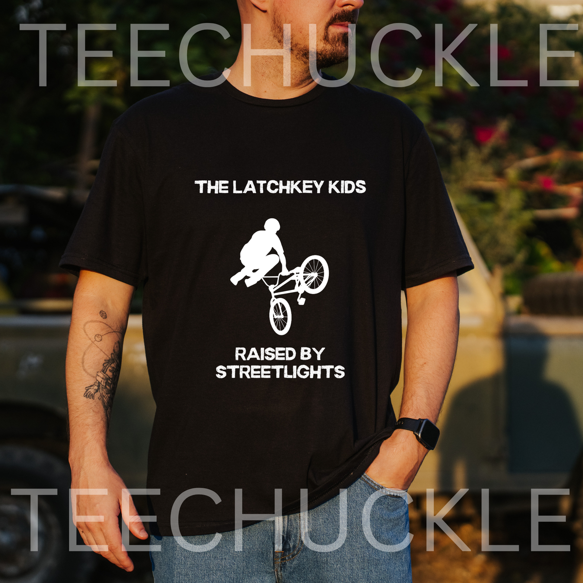 A man wearing a black graphic t-shirt with a white bicycle design and the text 'THE LATCHKEY KIDS RAISED BY STREETLIGHTS'. The background is an outdoor setting with a blurred vehicle and foliagE.