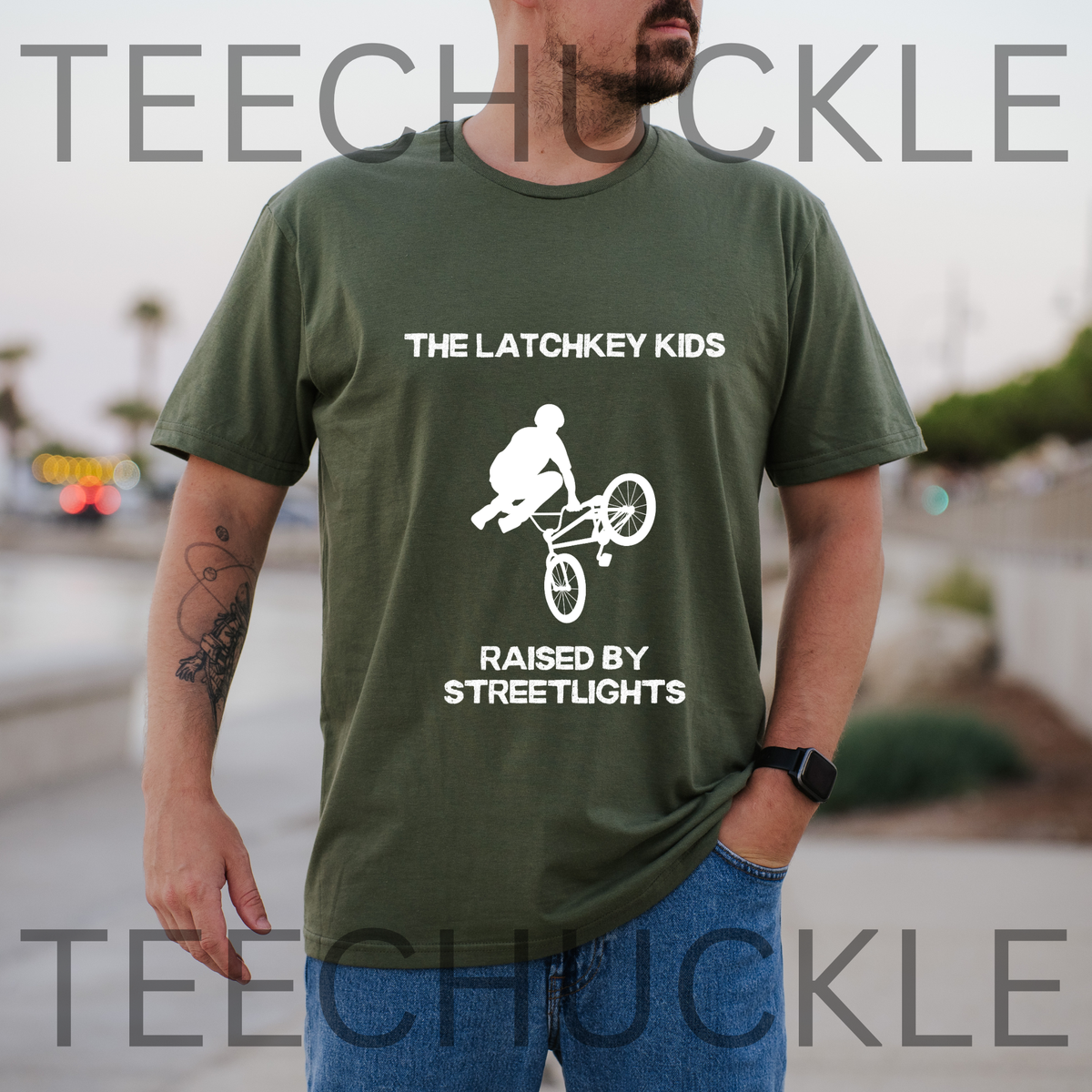 A man wearing a green graphic t-shirt with a white bicycle stunt print and the text 'THE LATCHKEY KIDS RAISED BY STREETLIGHTS, and the background is a blurred outdoor street scene.