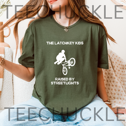 A woman wearing a green graphic t-shirt with a white bicycle graphic and the text 'THE LATCHKEY KIDS RAISED BY STREETLIGHTS'. 