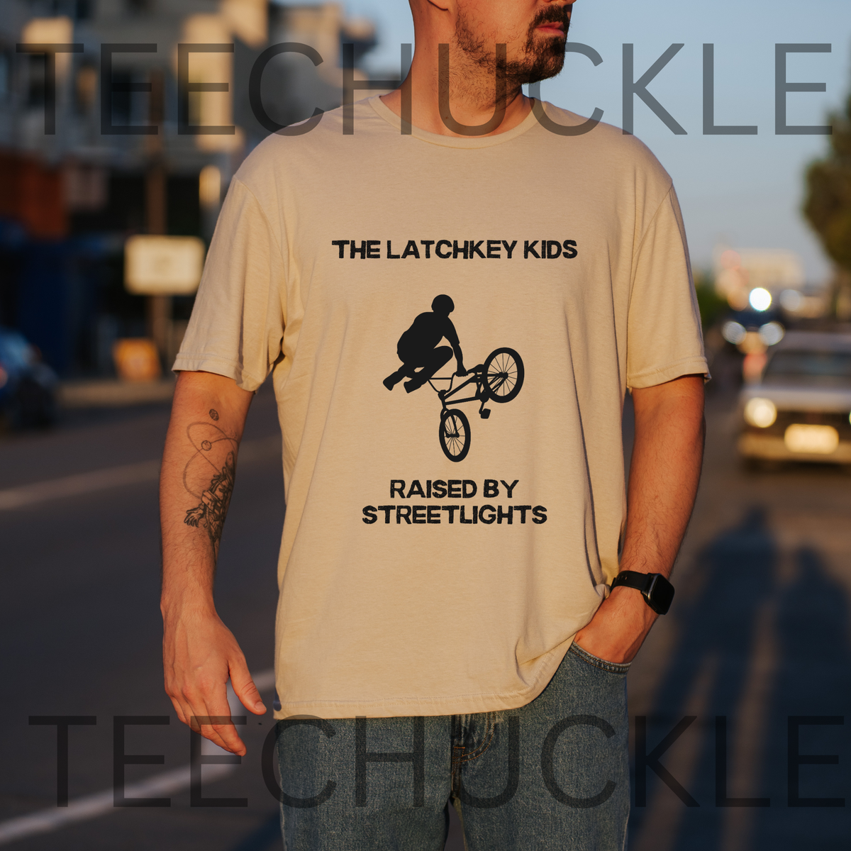 A man wearing a sand graphic t-shirt with a black bicycle stunt print and the text 'THE LATCHKEY KIDS RAISED BY STREETLIGHTS'. The photo is taken outdoors on a street at sunset