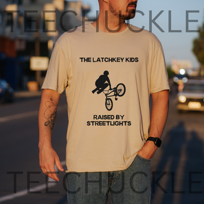 A man wearing a sand graphic t-shirt with a black bicycle stunt print and the text 'THE LATCHKEY KIDS RAISED BY STREETLIGHTS'. The photo is taken outdoors on a street at sunset
