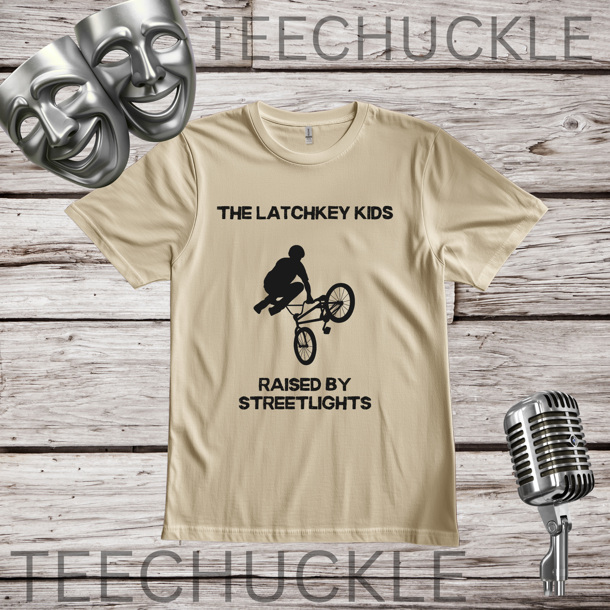 A sand graphic t-shirt displayed on a rustic wood background, featuring the black printed text 'THE LATCHKEY KIDS RAISED BY STREETLIGHTS' with a bicycle stunt graphic. The shirt is styled with silver comedy and tragedy masks, a vintage microphone.