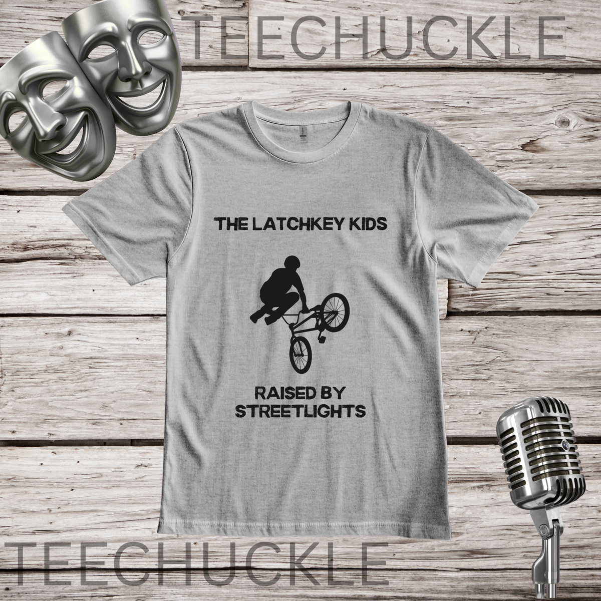 A gray graphic t-shirt displayed on a rustic wood background, featuring a black silhouette of a BMX rider with the text 'THE LATCHKEY KIDS RAISED BY STREETLIGHTS'. The shirt is styled with silver comedy masks, a vintage microphone