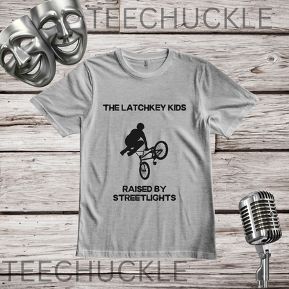 A gray graphic t-shirt displayed on a rustic wood background, featuring a black silhouette of a BMX rider with the text 'THE LATCHKEY KIDS RAISED BY STREETLIGHTS'. The shirt is styled with silver comedy masks, a vintage microphone
