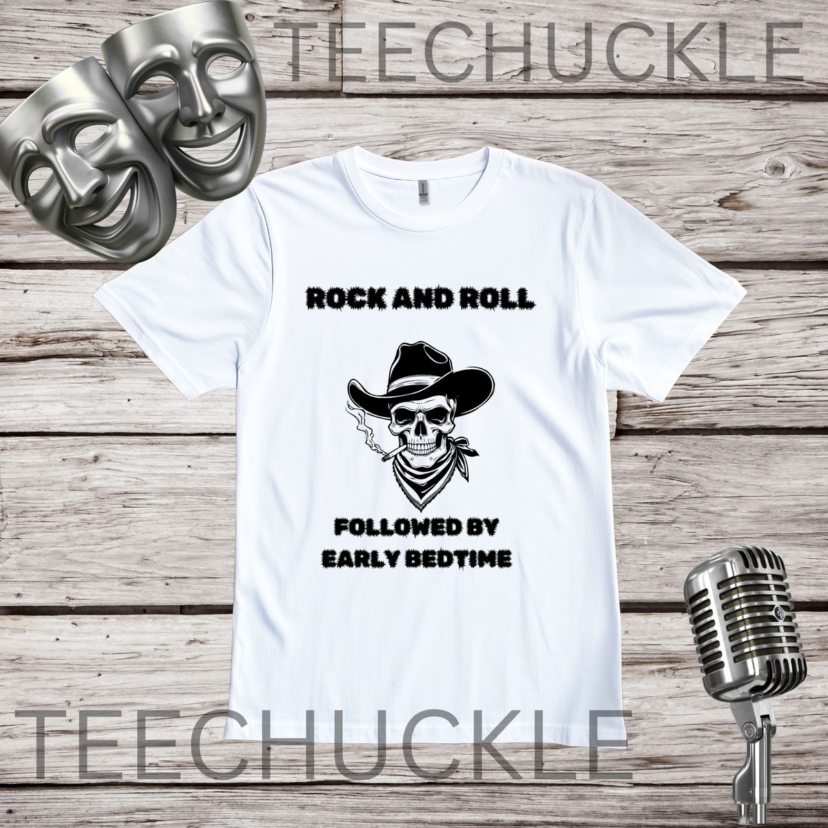 A white short-sleeve graphic T-shirt laid flat on a rustic wood plank background, with black text reading 'ROCK AND ROLL' above a skull wearing a cowboy hat and 'FOLLOWED BY EARLY BEDTIME' below. Two silver comedy and tragedy masks appear at top left, and a vintage microphone appears at bottom right.