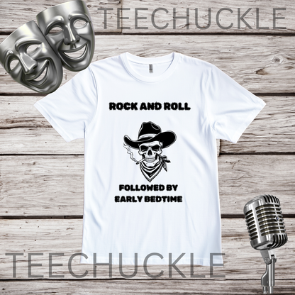 A white short-sleeve graphic T-shirt laid flat on a rustic wood plank background, with black text reading 'ROCK AND ROLL' above a skull wearing a cowboy hat and 'FOLLOWED BY EARLY BEDTIME' below. Two silver comedy and tragedy masks appear at top left, and a vintage microphone appears at bottom right.