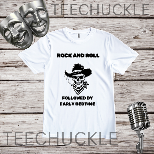 A white short-sleeve graphic T-shirt laid flat on a rustic wood plank background, with black text reading 'ROCK AND ROLL' above a skull wearing a cowboy hat and 'FOLLOWED BY EARLY BEDTIME' below. Two silver comedy and tragedy masks appear at top left, and a vintage microphone appears at bottom right.