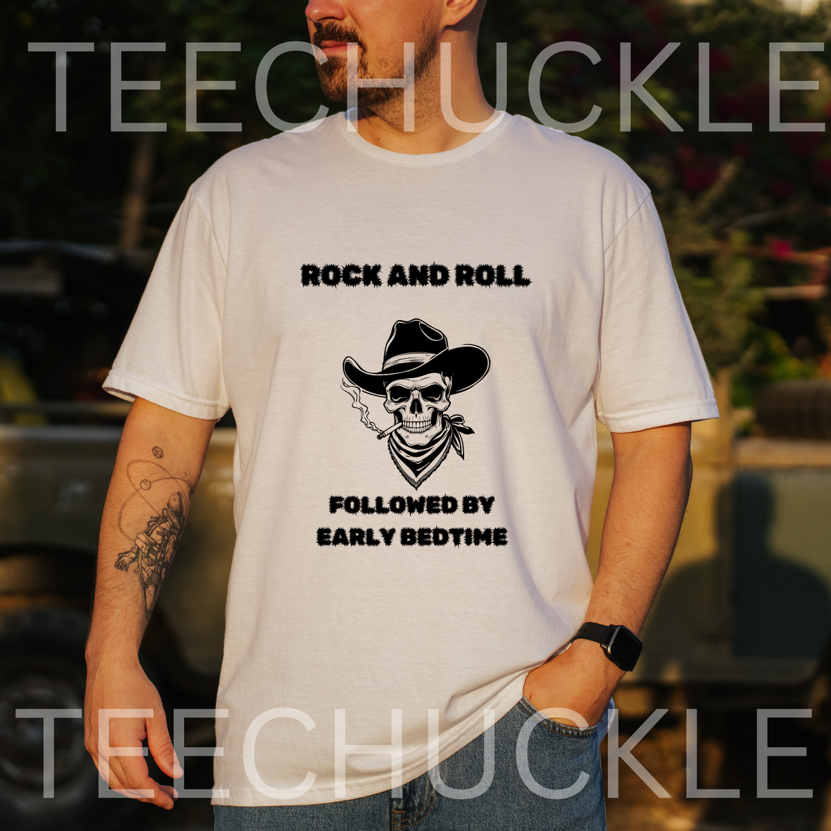 A man wearing a white graphic t-shirt with a black skull and cowboy hat design and the text 'ROCK AND ROLL FOLLOWED BY EARLY BEDTIME'. The image has a warm sunset lighting, a blurred outdoor background