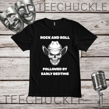 A black short-sleeve graphic T-shirt laid flat on a rustic wood plank background, with two silver theater masks, a vintage microphone. The shirt graphic shows skull with a cowboy hat on with the words “rock and roll, followed by an  early bedtime” in white text.