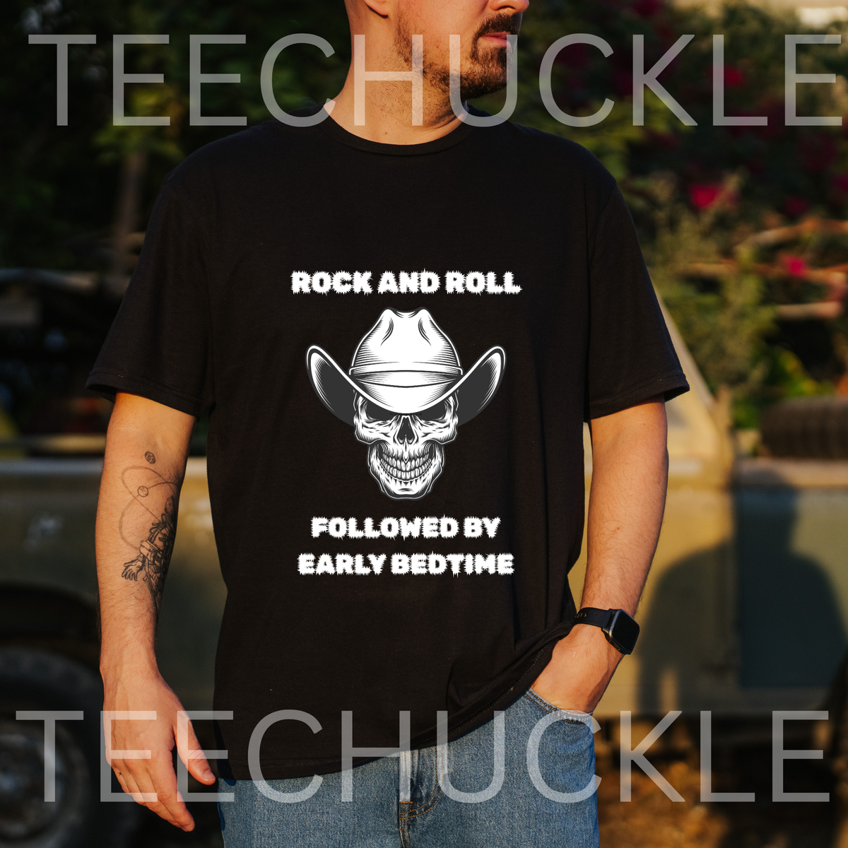 A man wearing a black graphic t-shirt with a white skull wearing a cowboy hat and the text 'ROCK AND ROLL FOLLOWED BY EARLY BEDTIME'. The background is an outdoor blurred setting with a vehicle and foliage.