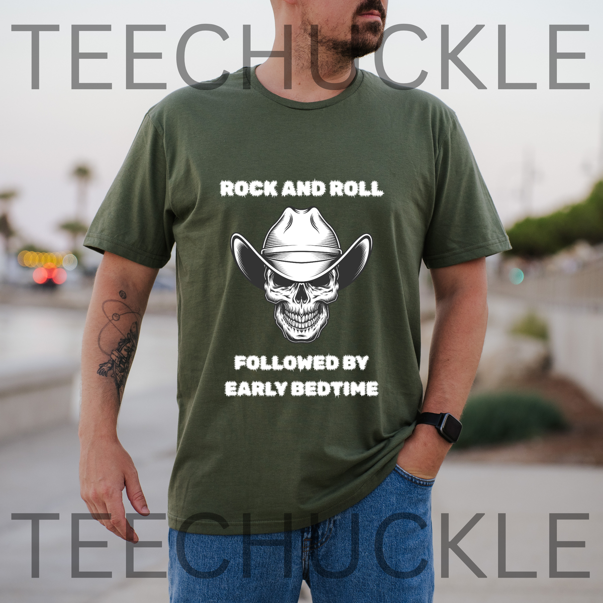 A man wearing an green graphic t-shirt with a white skull wearing a cowboy hat and the text 'ROCK AND ROLL FOLLOWED BY EARLY BEDTIME', and the background is a blurred outdoor street scene.