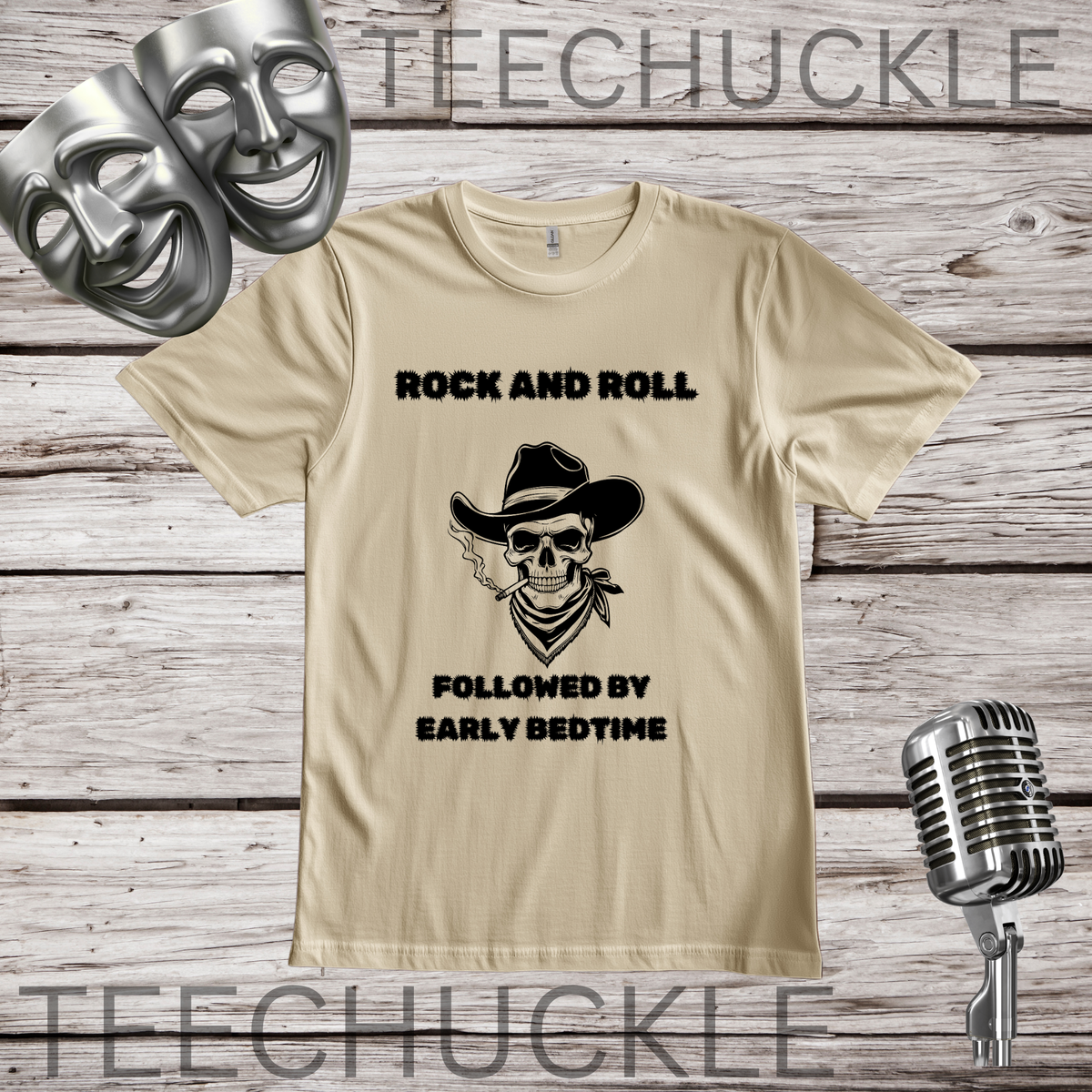 A sand graphic t-shirt displayed on a rustic wood background, featuring a black printed design of a cowboy skull and the text 'ROCK AND ROLL FOLLOWED BY EARLY BEDTIME'. The shirt is styled with silver comedy and tragedy masks, a vintage microphone.