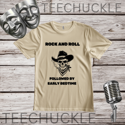 A sand graphic t-shirt displayed on a rustic wood background, featuring a black printed design of a cowboy skull and the text 'ROCK AND ROLL FOLLOWED BY EARLY BEDTIME'. The shirt is styled with silver comedy and tragedy masks, a vintage microphone.