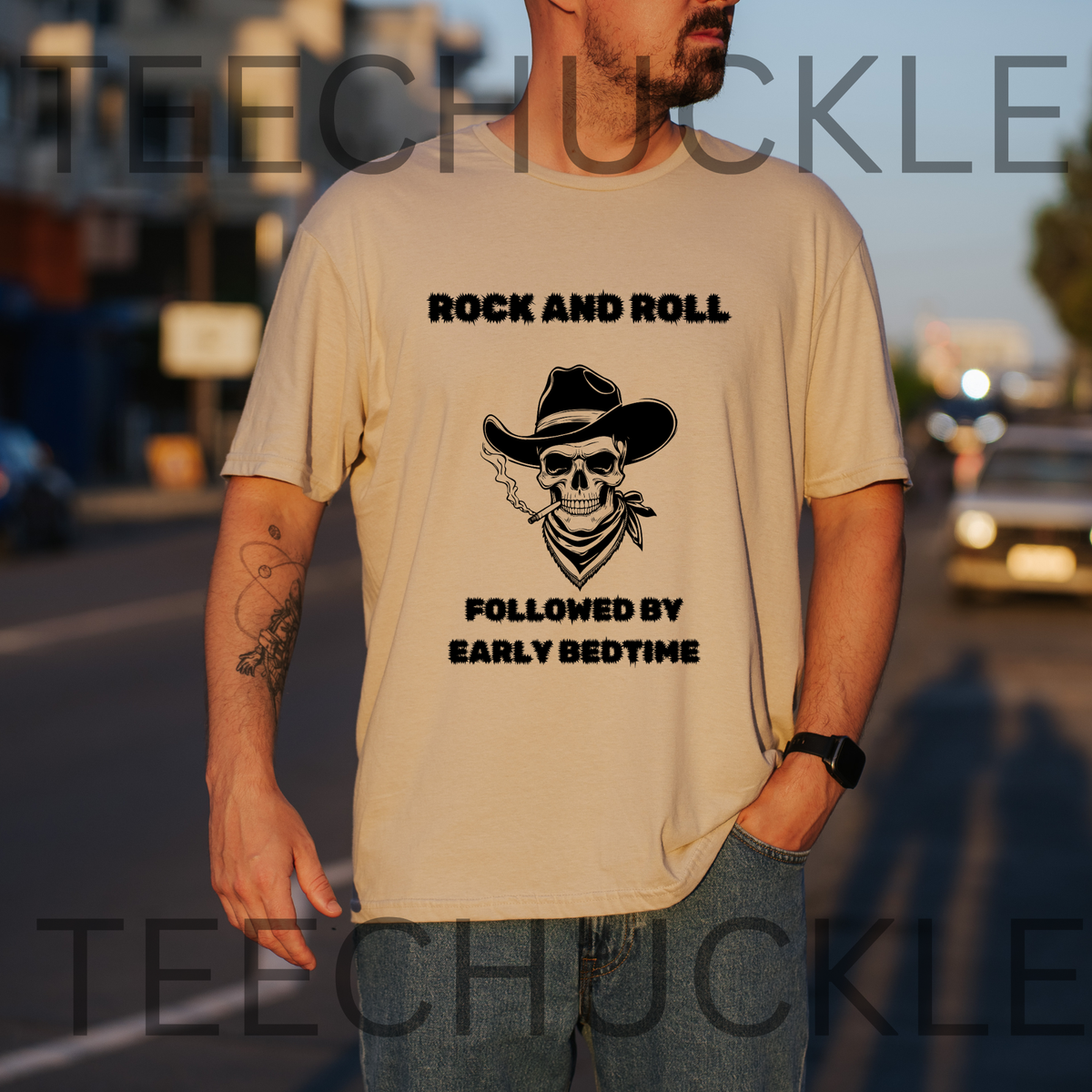 A man wearing a sand graphic t-shirt with a black skull and cowboy hat design and the text 'ROCK AND ROLL FOLLOWED BY EARLY BEDTIME'. The background is a blurred street at sunset.