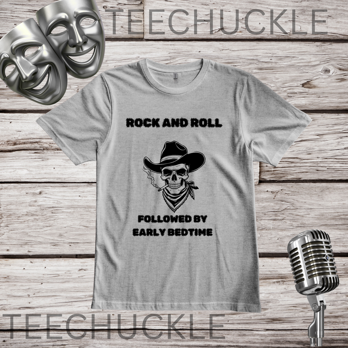 A light gray graphic t-shirt featuring a black printed design of a cowboy skull with a bandana and the text 'ROCK AND ROLL FOLLOWED BY EARLY BEDTIME'. The t-shirt is displayed on a rustic wood background with a vintage microphone, comedy masks.