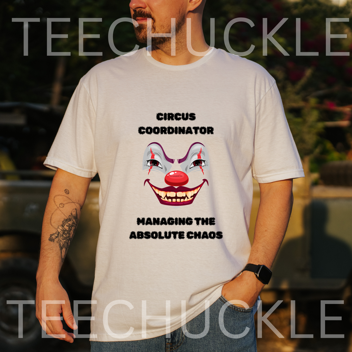 A man wearing a white graphic t-shirt with a clown face design and the text 'CIRCUS COORDINATOR' and 'MANAGING THE ABSOLUTE CHAOS'. The background is a blurred outdoor setting with a watermark 'TEECHUCKLE' across the image.