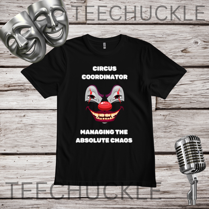 A black short-sleeve graphic t-shirt displayed on a rustic wood background with theatrical props. The shirt features a clown face graphic with red lips and white text reading 'CIRCUS COORDINATOR' and 'MANAGING THE ABSOLUTE CHAOS'.