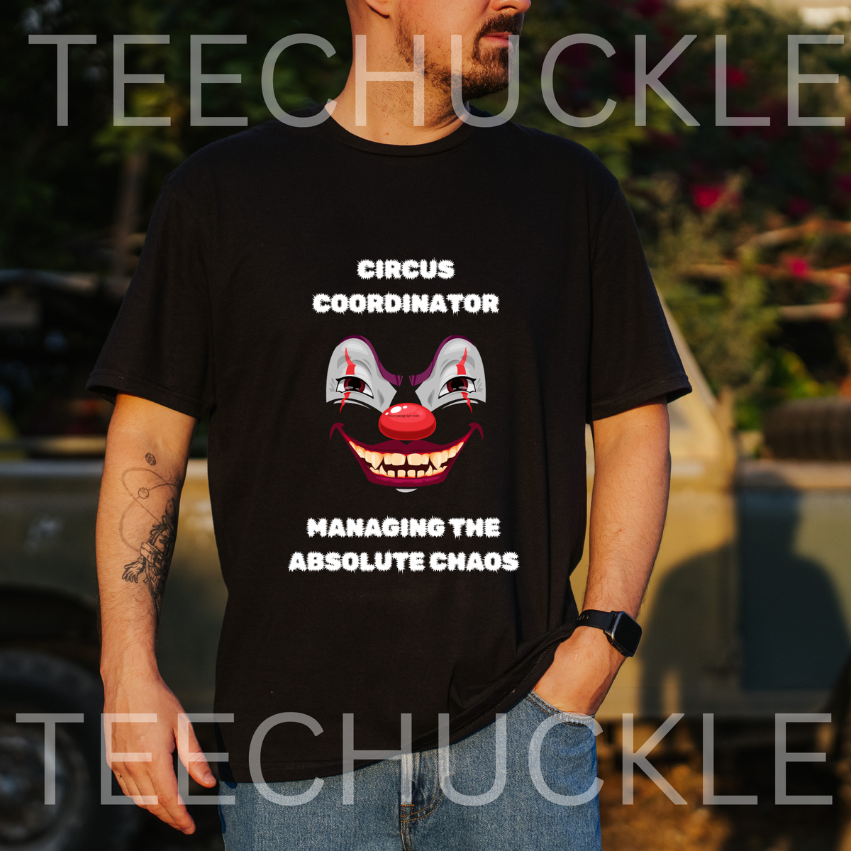 A man wearing a black graphic t-shirt with a clown face and the text 'CIRCUS COORDINATOR MANAGING THE ABSOLUTE CHAOS'. The background is a blurred outdoor setting