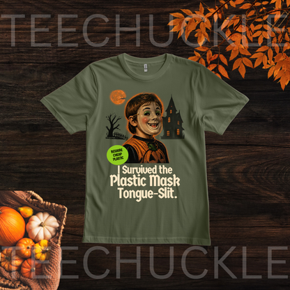A green graphic t-shirt displayed flat on a dark wood background with autumn-themed props like pumpkins and orange leaves. The graphic features a cartoon of a smiling child with a plastic mask and a tongue slit, with the text 'I Survived the Plastic Mask Tongue-Slit.