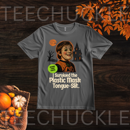 A dark gray graphic t-shirt displayed flat on a dark wood background with autumn-themed props like pumpkins and orange leaves. The graphic features a cartoon illustration of a smiling woman in a plastic mask with a haunted house and full moon in the background, accompanied by the text 'I Survived the Plastic Mask Tongue-Slit.