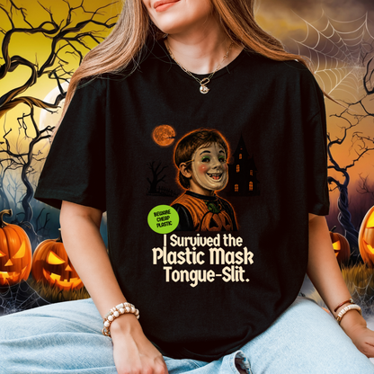 A woman wearing a black graphic t-shirt with a Halloween-themed illustration and the text 'I Survived the Plastic Mask Tongue-Slit.' She is sitting in front of a spooky Halloween background with pumpkins, cobwebs, and a haunted house.