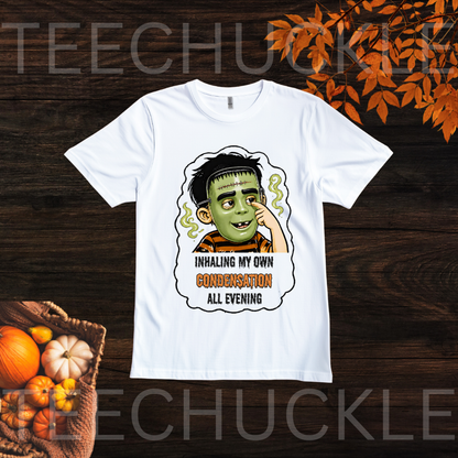 A white short-sleeve graphic T-shirt laid flat on a dark wood background with autumn leaves and pumpkins. The shirt features a cartoon child with a halloween mask on character and the text 'INHALING MY OWN CONDENSATION ALL EVENING.