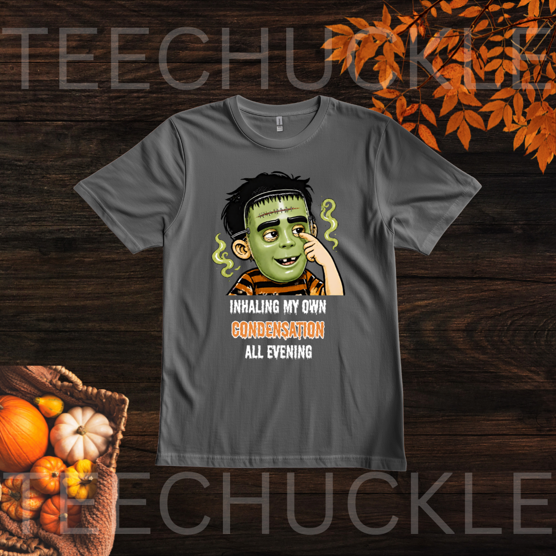 A dark gray graphic t-shirt featuring a cartoon illustration of a child wearing a halloween mask with the text 'INHALING MY OWN CONDEMNATION ALL EVENING'. The t-shirt is displayed on a dark wood background with Halloween-themed props including pumpkins and autumn leaves