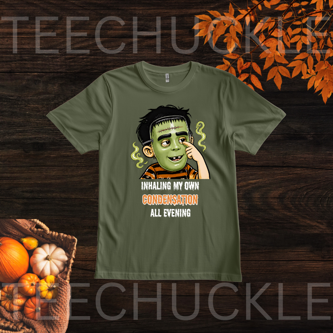 A green graphic t-shirt featuring a cartoon illustration of a child in a halloween mask with the text 'INHALING MY OWN CONDENSATION ALL EVENING'. The t-shirt is displayed flat on a dark wood background with decorative autumn leaves and pumpkins in the corners.