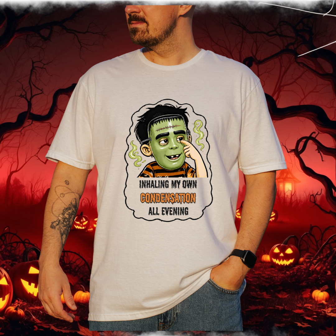 A man wearing a white graphic t-shirt with a cartoon illustration of a child in a halloween mask and the text 'INHALING MY OWN CONDEMNATION ALL EVENING'. The background is a spooky Halloween scene with pumpkins and bare trees.