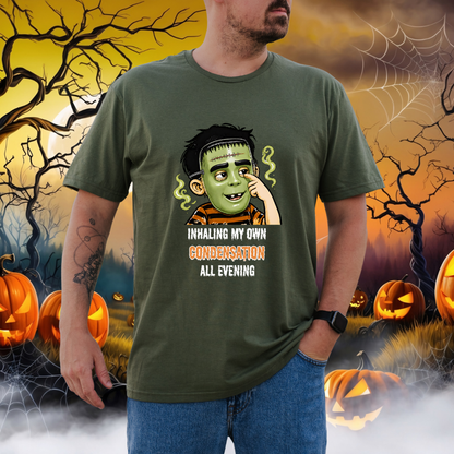 A man wearing an olive green graphic t-shirt with a cartoon child in a halloween mask and the text 'INHALING MY OWN CONDENSATION ALL EVENING'. The background is a spooky Halloween scene with jack-o'-lanterns, cobwebs, and a full moon.