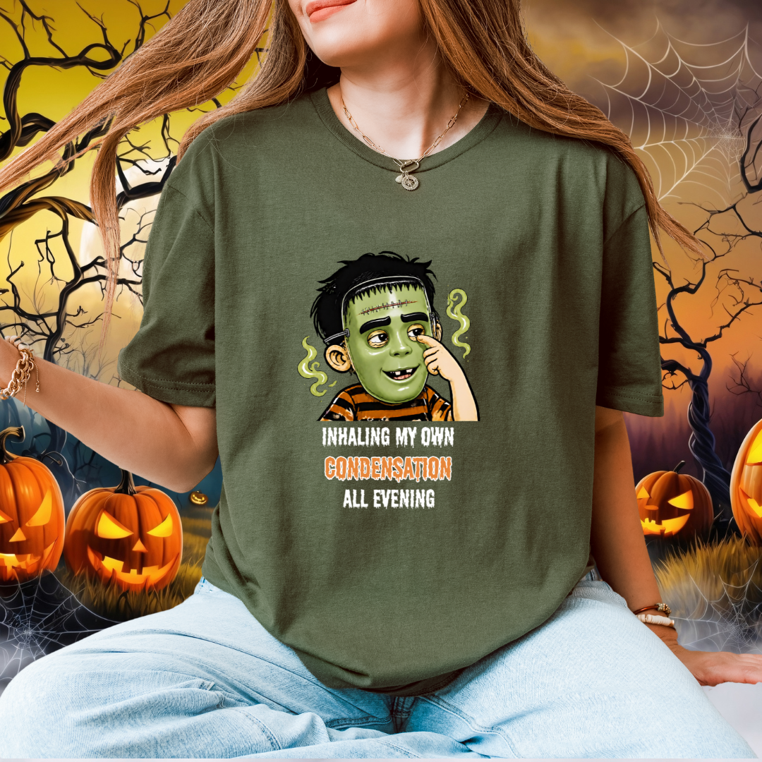 A woman wearing an olive green graphic t-shirt with a cartoon child with a halloween mask illustration and the text 'INHALING MY OWN CONDEMNATION ALL EVENING'. The background is a Halloween-themed digital scene with pumpkins, cobwebs, and a full moon.