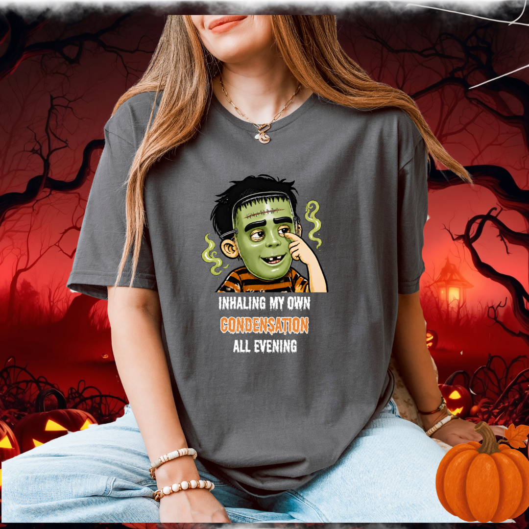 A woman wearing a dark gray graphic t-shirt with a cartoon character in a halloween mask and the text 'INHALING MY OWN CONDENSATION ALL EVENING'. The background is a spooky Halloween scene with pumpkins and a red sky.