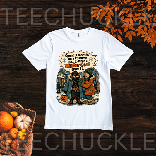 A white t-shirt laid flat on wood with autuum leaves and pumpkins around it. The shirt has a illustration of a child in ahalloween costume with the text "SPENT 3 MONTHS ON A COSTUME JUST TO WEAR A WINTER COAT OVER IT"