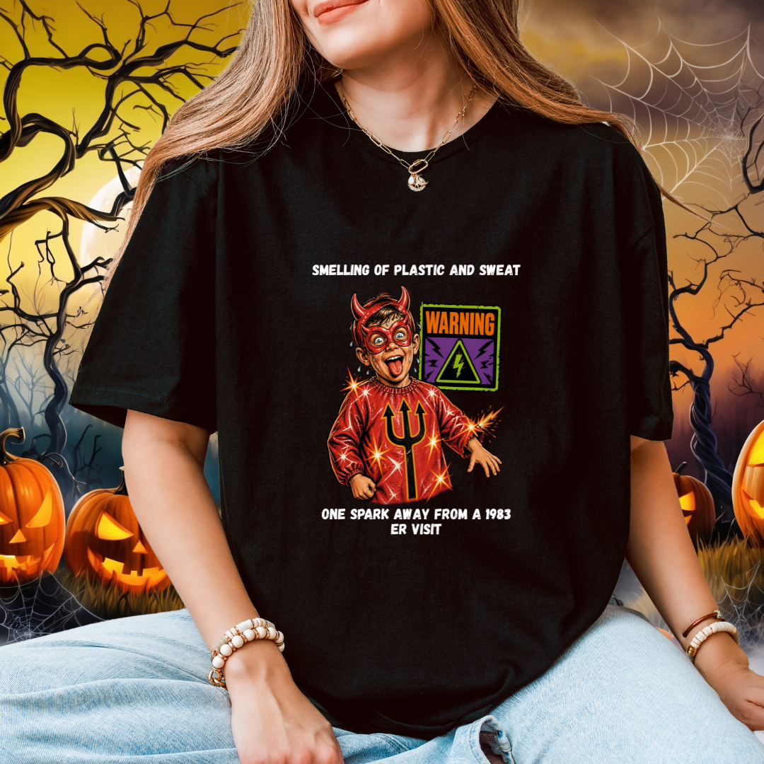 A woman wearing a black graphic t-shirt with a devil-themed print and humours text, posed against a spooky Halloween background with pumpkins and cobwebs.