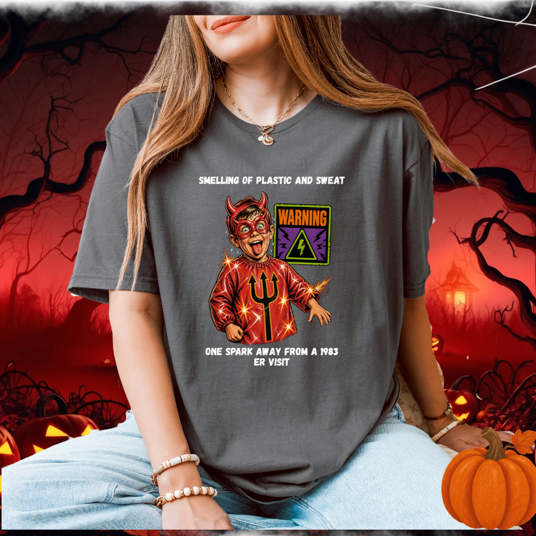 A woman wearing a dark gray graphic t-shirt with a devil-themed design with humourous text, posed in a spooky Halloween-themed setting with pumpkins and a red background.