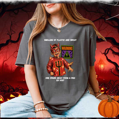 A woman wearing a dark gray graphic t-shirt with a devil-themed design with humourous text, posed in a spooky Halloween-themed setting with pumpkins and a red background.