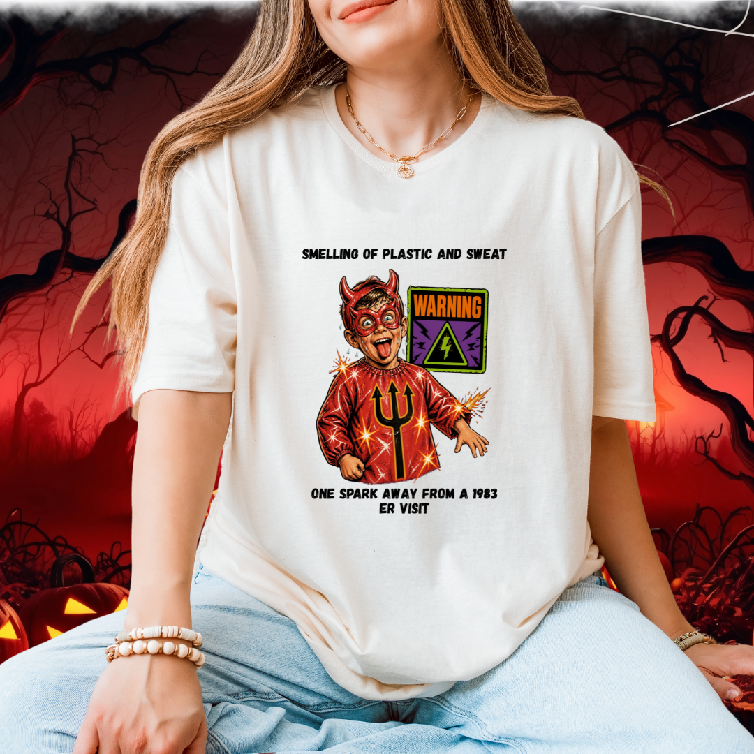 A woman wearing a white graphic t-shirt with a devil design and the text 'SMELLING OF PLASTIC AND SWEAT' and 'ONE SPARK AWAY FROM A 1983 ER VISIT'. The background is a spooky red and black Halloween scene with jack-o'-lanterns.