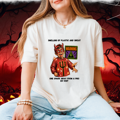 A woman wearing a white graphic t-shirt with a devil design and the text 'SMELLING OF PLASTIC AND SWEAT' and 'ONE SPARK AWAY FROM A 1983 ER VISIT'. The background is a spooky red and black Halloween scene with jack-o'-lanterns.