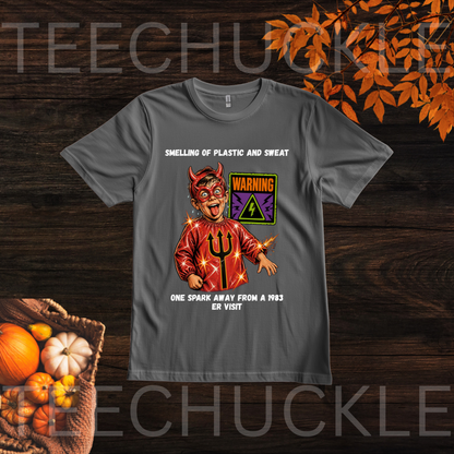 A dark gray graphic t-shirt displayed flat on a dark wood background with autumn-themed props like pumpkins and orange leaves. The t-shirt features a graphic of a devil character in a red suit with a trident, a warning sign, and humorous text.